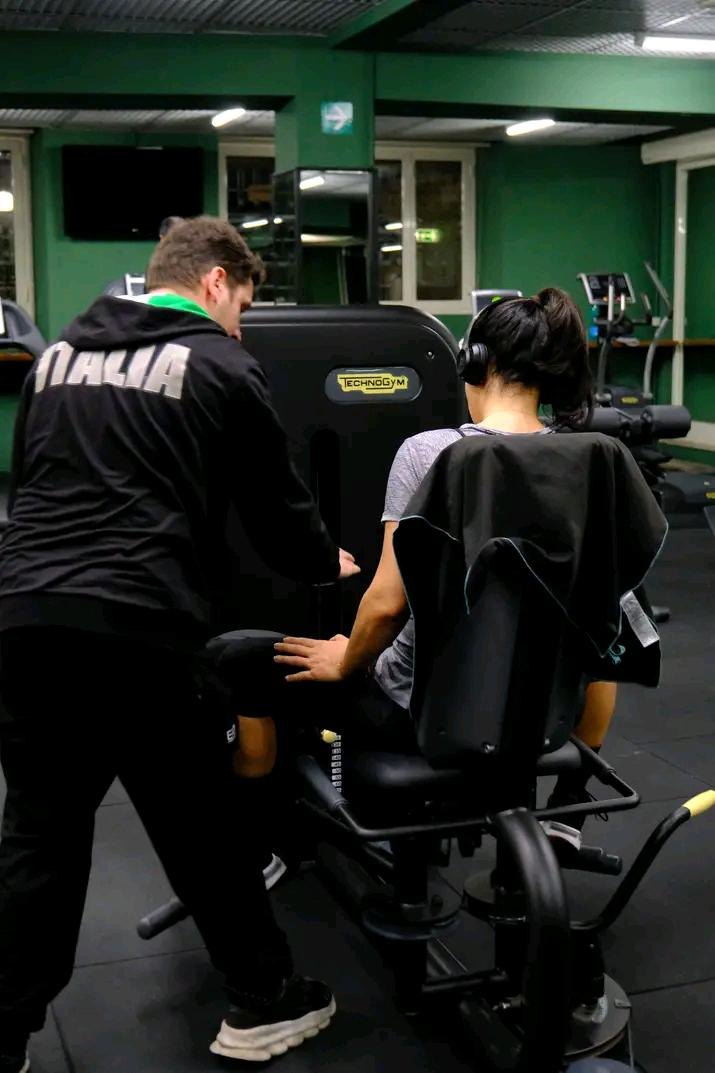 Trainer con felpa ITALIA assiste una donna con cuffie su macchina Technogym per le gambe, tra pareti verdi