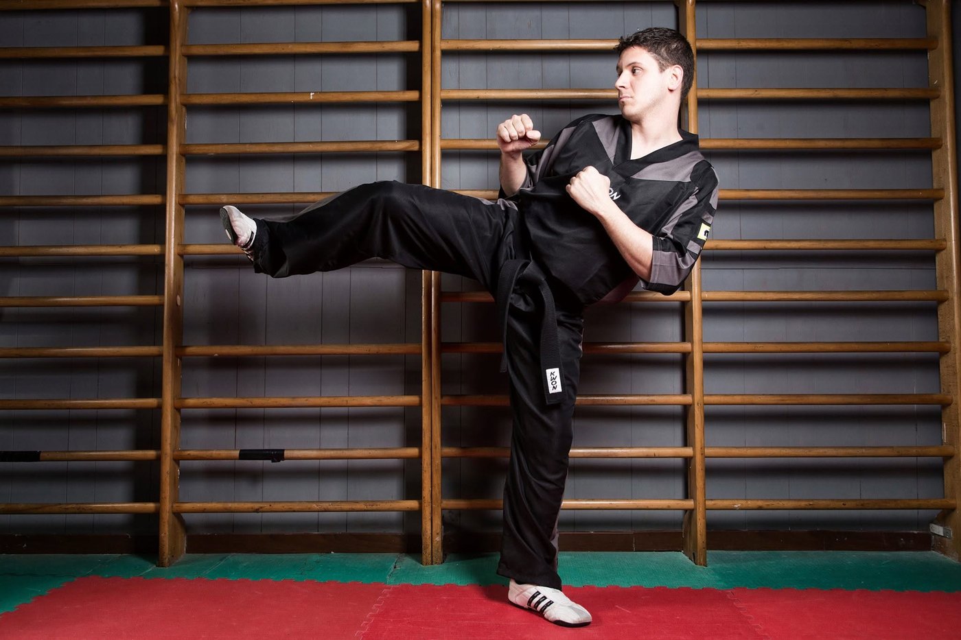 Uomo in divisa nera e cintura nera esegue un calcio laterale davanti a spalliere di legno su tatami rosso e verde